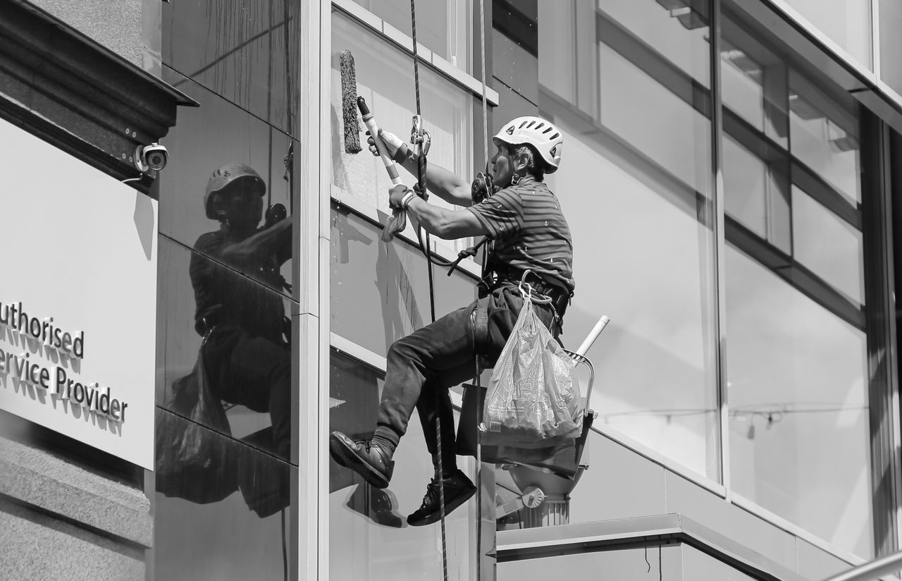 services-02 Black and white, viewed from below, of a steeplejack wearing a protective helmet and professional gear, cleaning the glass facade of a modern tower